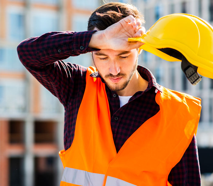 An exhausted worker in an orange safety vest holding a yellow hard hat near his forehead.