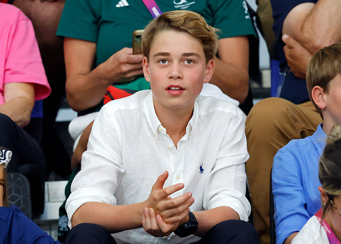 Prince George wearing a white collared shirt and looking ahead while sitting in a crowd. Prince George wearing a white collared shirt and looking ahead while sitting in a crowd.