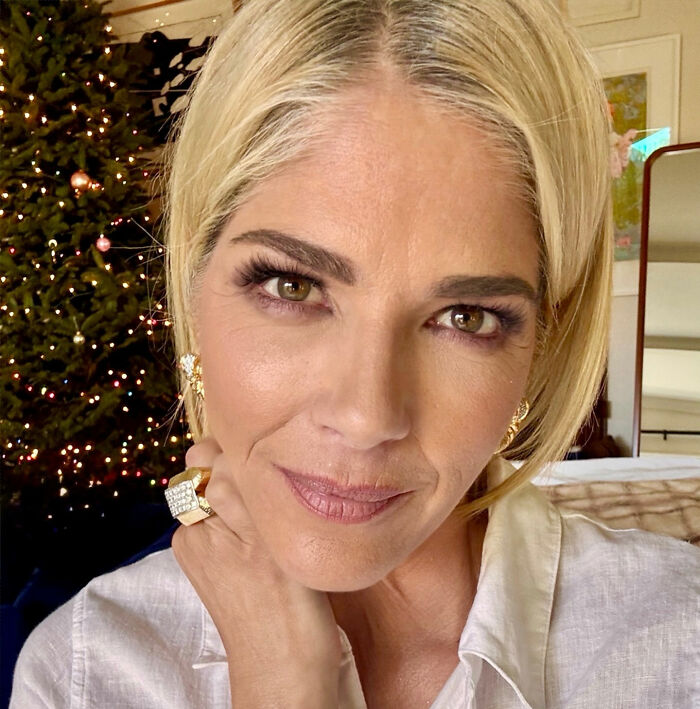 Selma Blair posing for a close-up selfie with blonde hair and gold jewelry near a decorated Christmas tree.