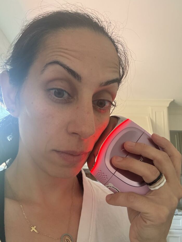 A woman demonstrating a pink red-light therapy device on her face.