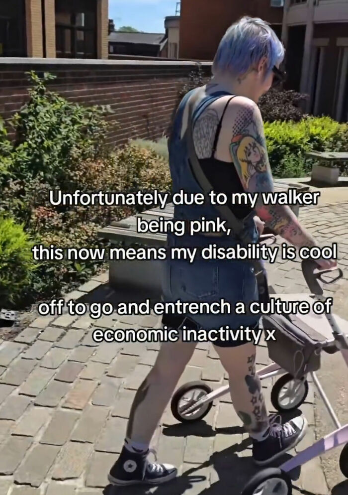 A tattooed young woman with blue hair walking outdoors while using a pink walker mobility aid.