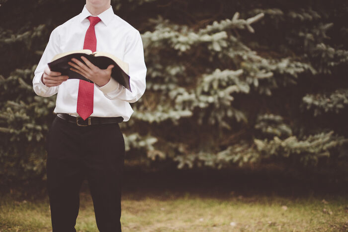 A man in a white shirt and red tie holding an open book outdoors, reflecting spiritual cults
