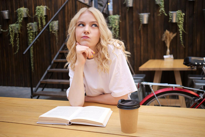 A blonde girl sitting thoughtful with a book and coffee cup, contemplating school and honest fundraiser concepts.