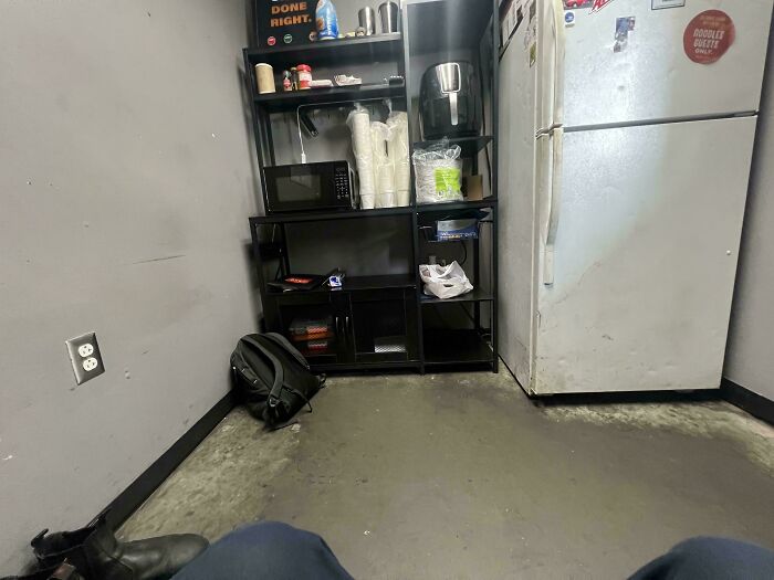 A cramped breakroom with basic appliances and a backpack on the floor, capturing an aspect of modern life in America.