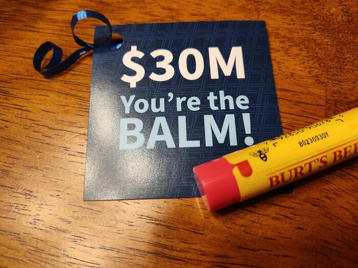 A blue gift tag reading $30M You are the BALM next to a lip balm on a wooden surface, showing modern life in America.