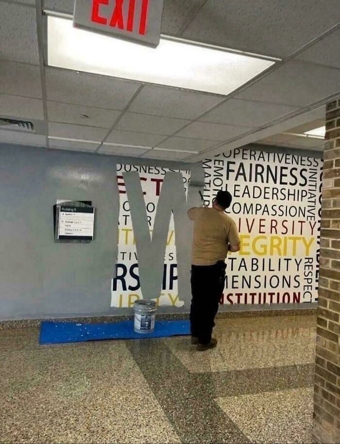 A painter covering up a school wall mural with positive words, illustrating aspects of modern life in America.