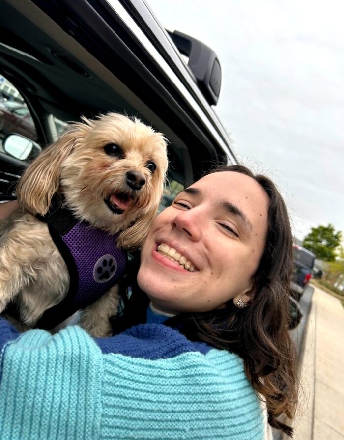 Smiling woman takes a selfie in a car with her small, fluffy dog, celebrating a viral TikTok trend about pets.
