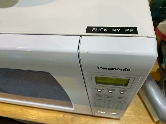 Vandalism on a white microwave with label maker tape reading SUCK MY PP
