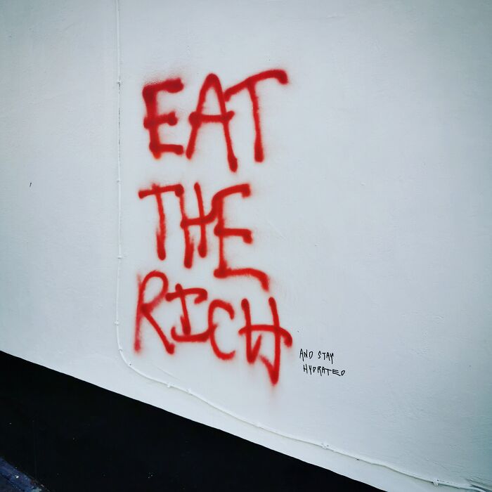Red graffiti on a wall reading Eat the Rich with clever vandalism adding and stay hydrated.