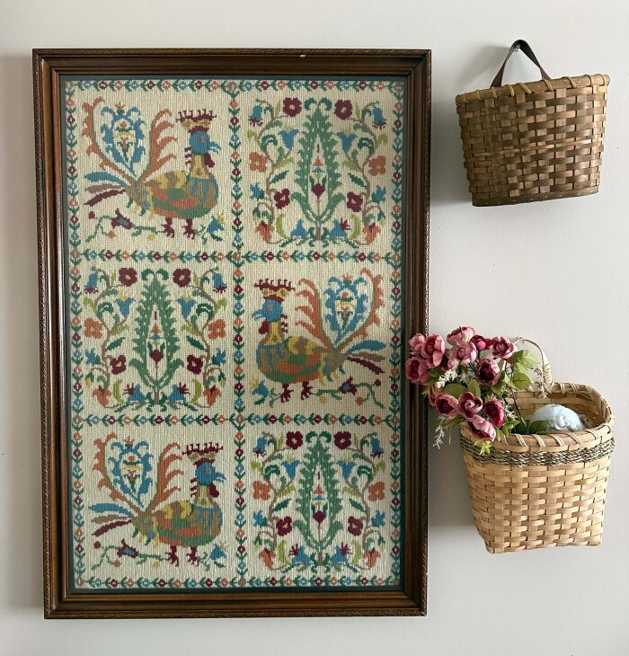 A framed tapestry with rooster designs hung next to small baskets, representing unique thrift store finds