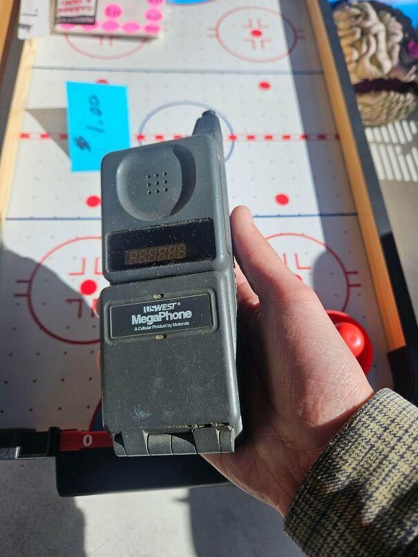 A hand holding a thick, blocky grey vintage tech mobile phone over an air hockey table