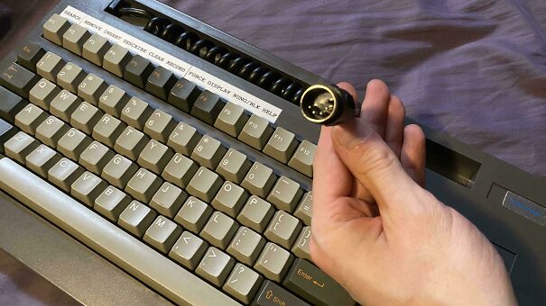A hand holding a vintage tech keyboard cable next to its retro chunky keyboard