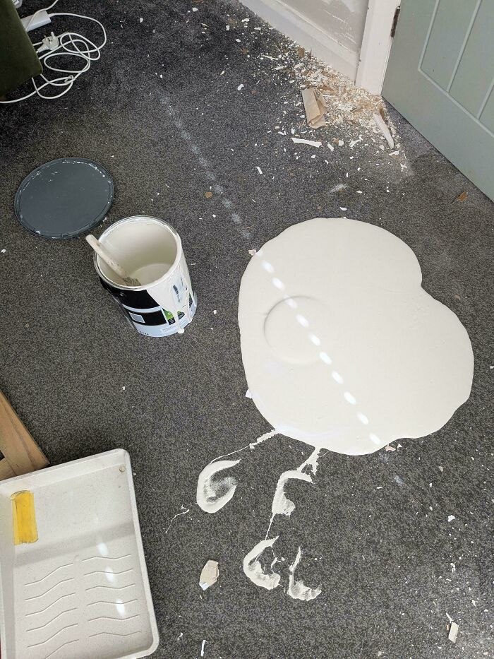 A questionable decision showing white paint spilled on a grey carpeted floor with footprints nearby