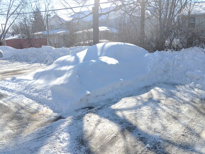 A car completely buried under a thick, smooth layer of white snow in a parking lot, highlighting questionable decisions.