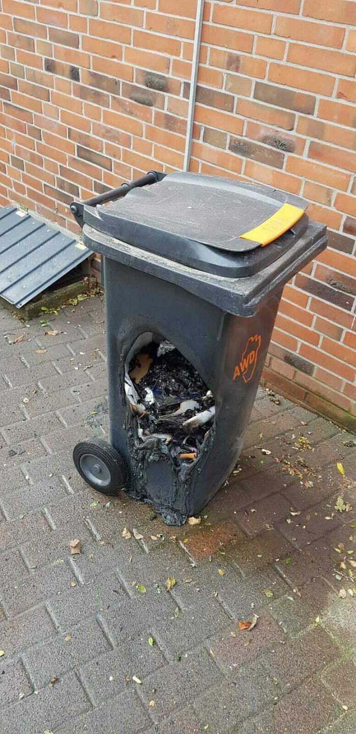 A melted black trash can with a large hole in the side from a hot coal, showing questionable decisions