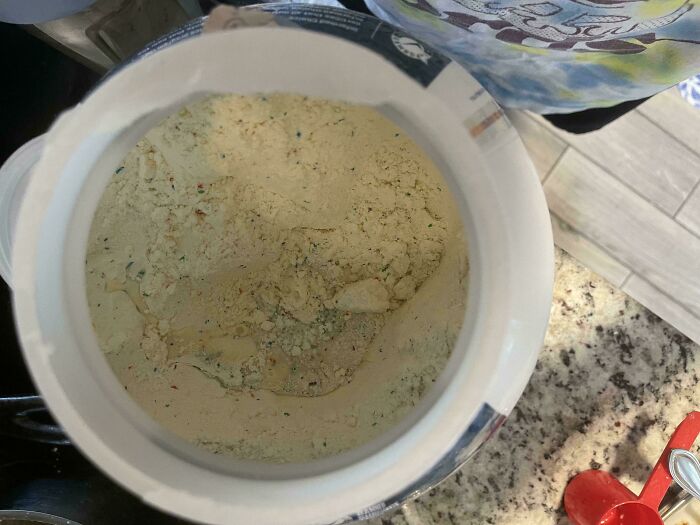 Opened protein powder tub containing a layer of damp, discolored clumps on the surface