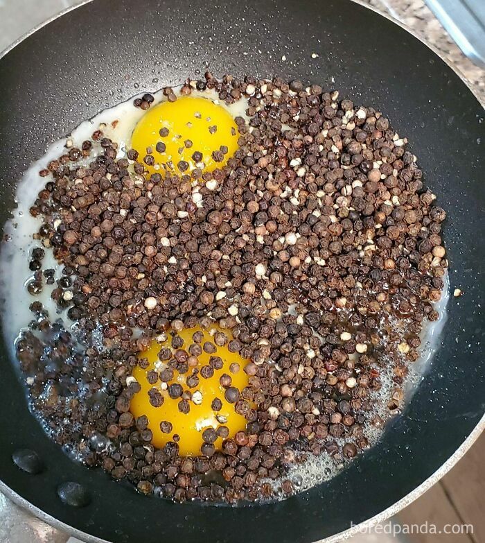 Two eggs frying in a pan covered completely in whole black peppercorns, a questionable decision