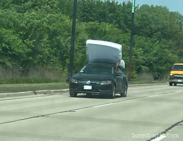 A black sedan driving down the highway with a mattress on its roof held only by the driver's hand.