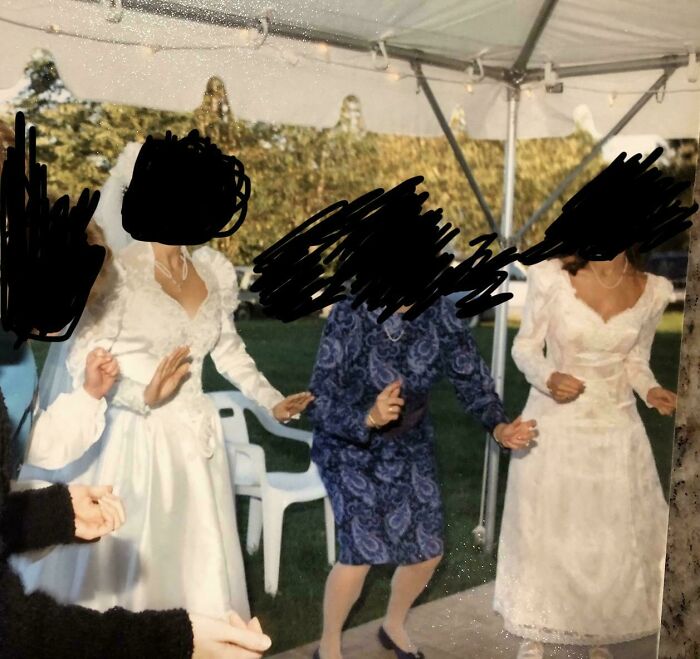 A questionable wedding moment of three women in white gowns holding hands and dancing under a tent