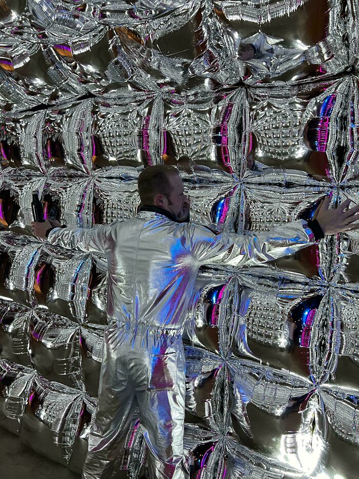 A man in a silver jumpsuit blending into a wall of silver foil balloons as accidental camouflage