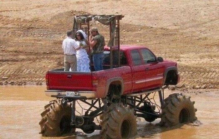 A couple having a questionable wedding ceremony standing in the bed of a monster truck parked in mud.