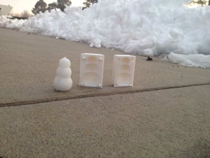 A small snowman next to a two-part 3D printer mold on a concrete sidewalk, showcasing human ingenuity.