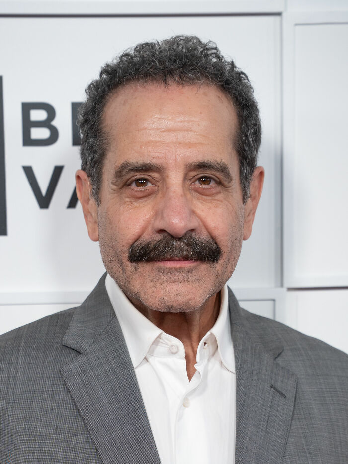 A close-up portrait of actor Tony Shalhoub smiling in a grey suit jacket and white collared shirt.
