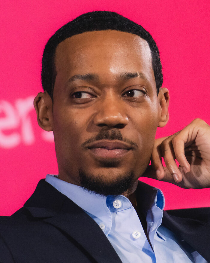 A close-up portrait of Tyler James Williams posing in front of a vibrant pink background.