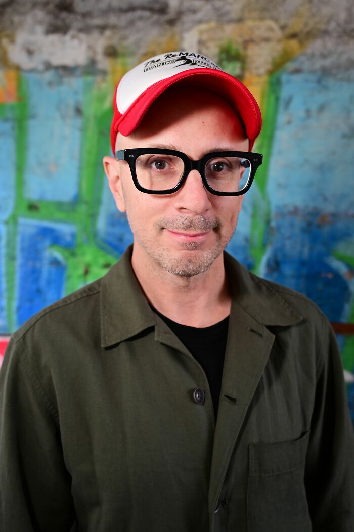 Steve Burns wearing a red cap and black glasses posing in front of a colorful graffiti wall.