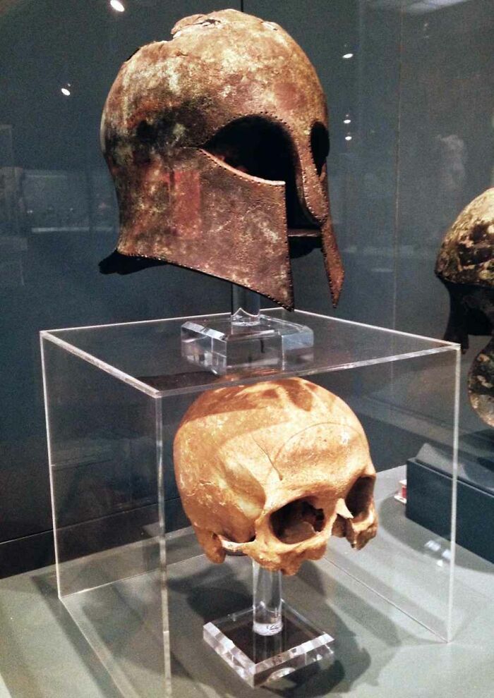 Rusty ancient helmet displayed above old human skull in museum