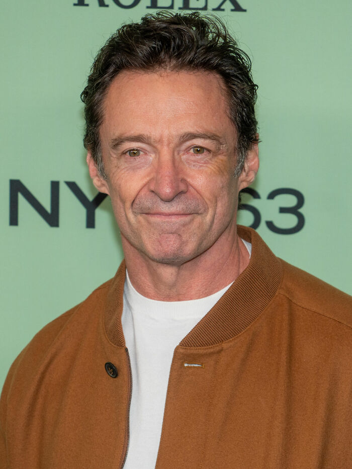Actor Hugh Jackman smiling at a red carpet event in a brown jacket