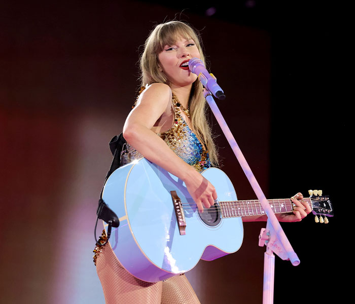 Taylor Swift playing a light blue acoustic guitar during a live stage performance.