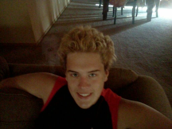 Blonde man sitting on a couch smiling in a grainy low-light setting, reminiscent of creepy photos.