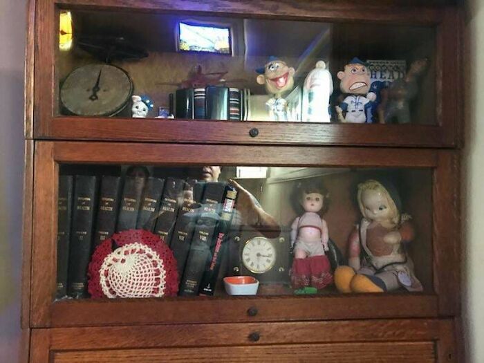 Creepy photos of vintage dolls and knickknacks displayed inside a glass cabinet with an unsettling reflection.