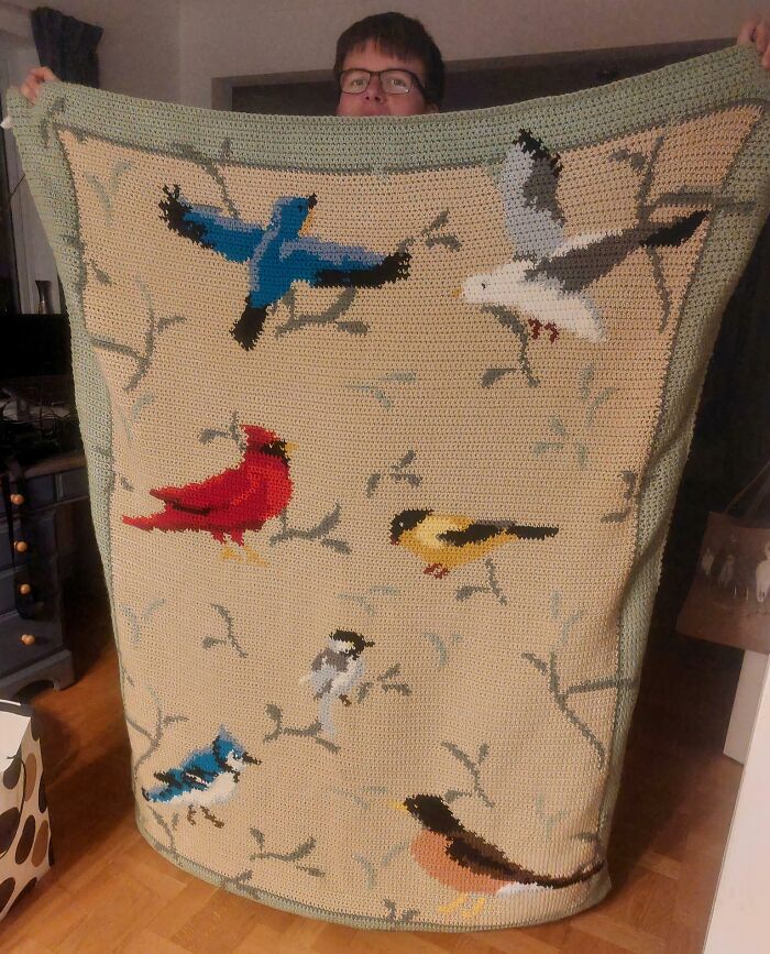 Colorful bird pattern crochet blanket held up by person indoors