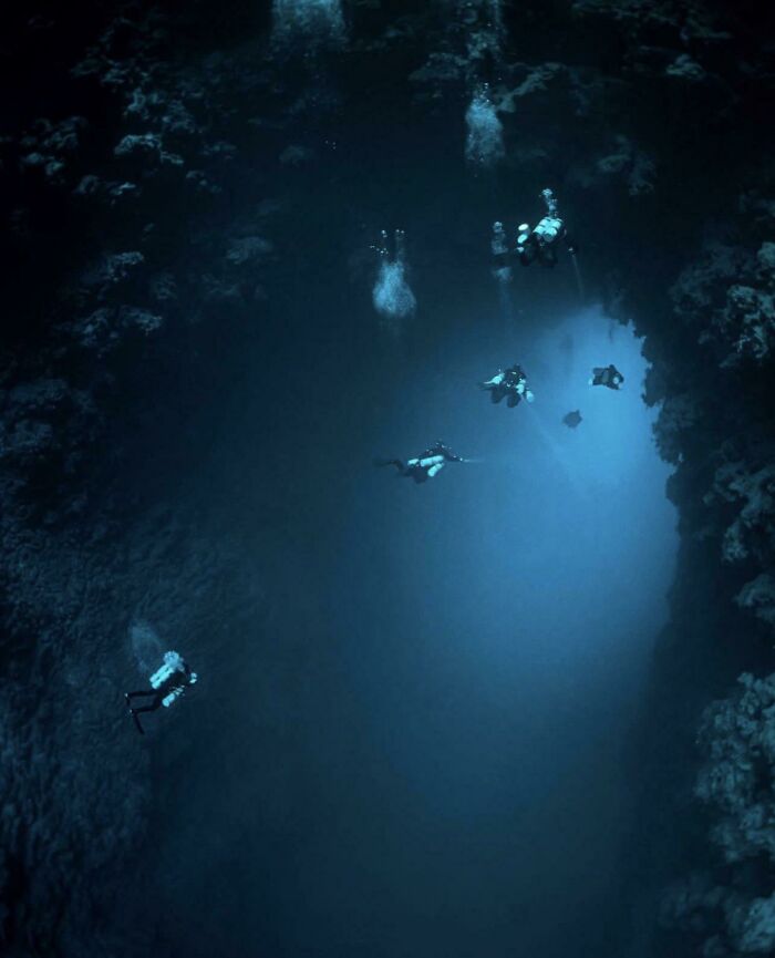 Group of divers descending into a dark deep-water underwater cave with blue light