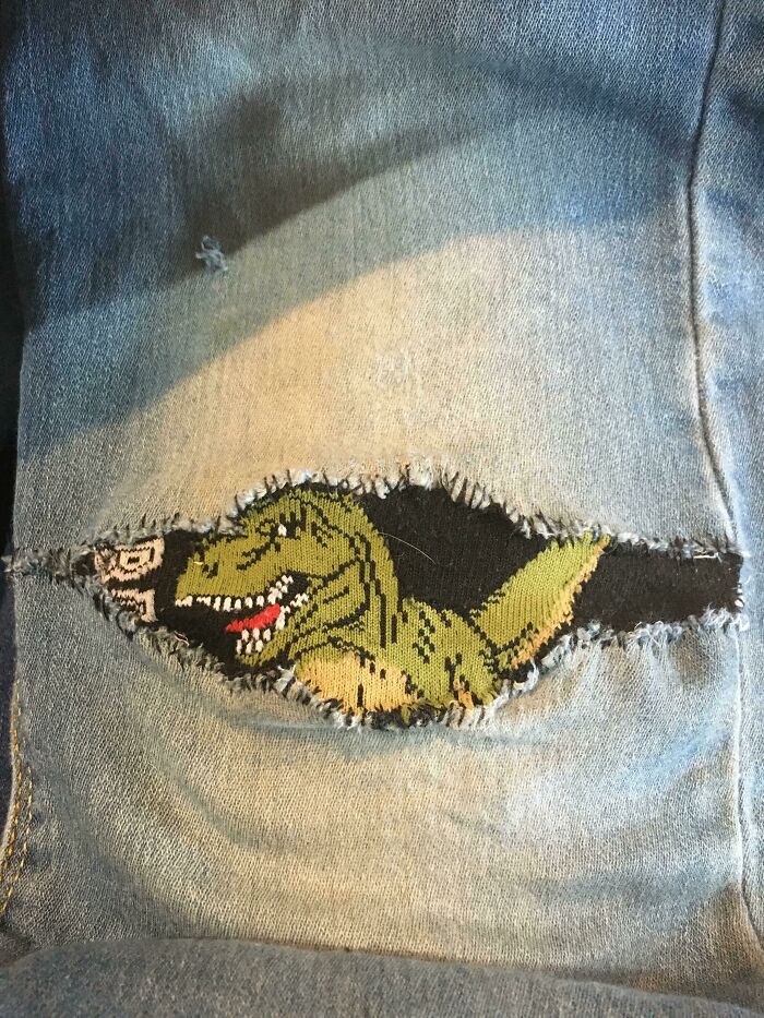 A green dinosaur patch peeking through a tear in blue jeans, showing creativity to mend clothes and save money.
