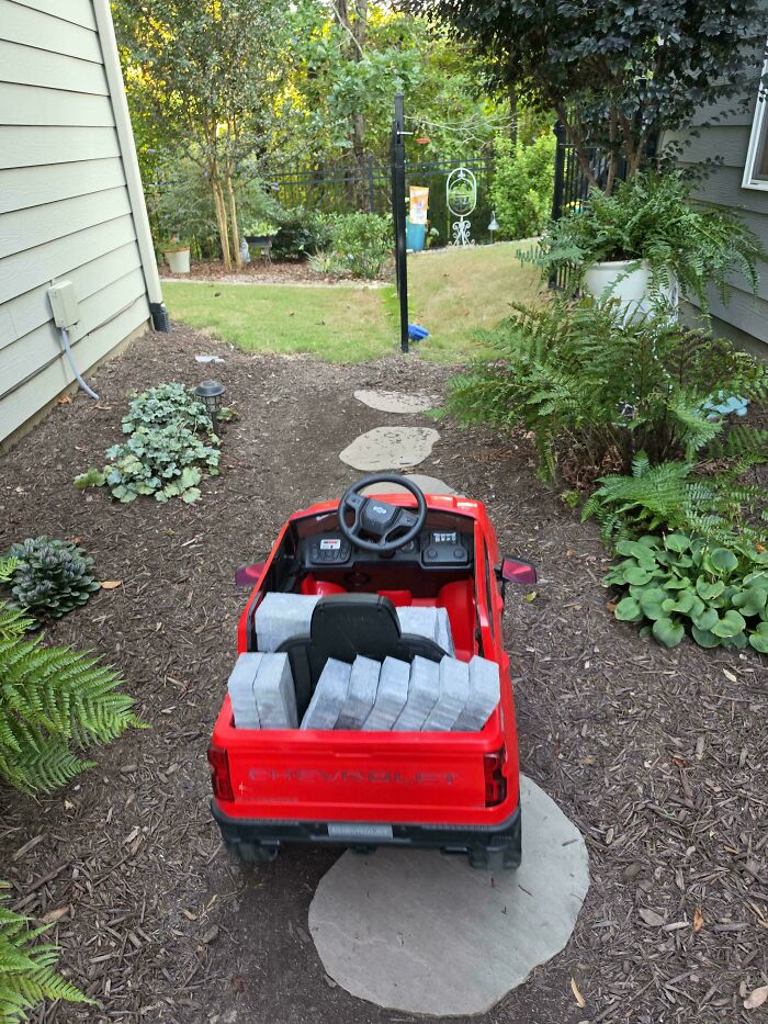 Red toy ride-on truck loaded with pavers in a garden path created by hardworking dads.