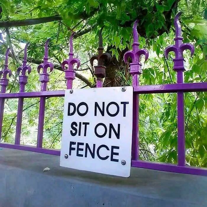 A sign on a sharp purple metal fence displaying confusing instructions that say do not sit on fence.