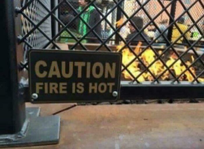 A sign on a metal grid fence showing hilariously confusing labels stating caution fire is hot.
