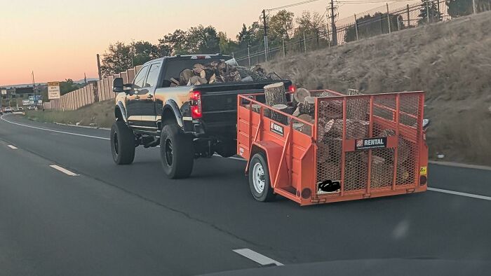 A black truck towing an orange rental trailer overflowing with heavy logs, illustrating an example of towing gone wrong.