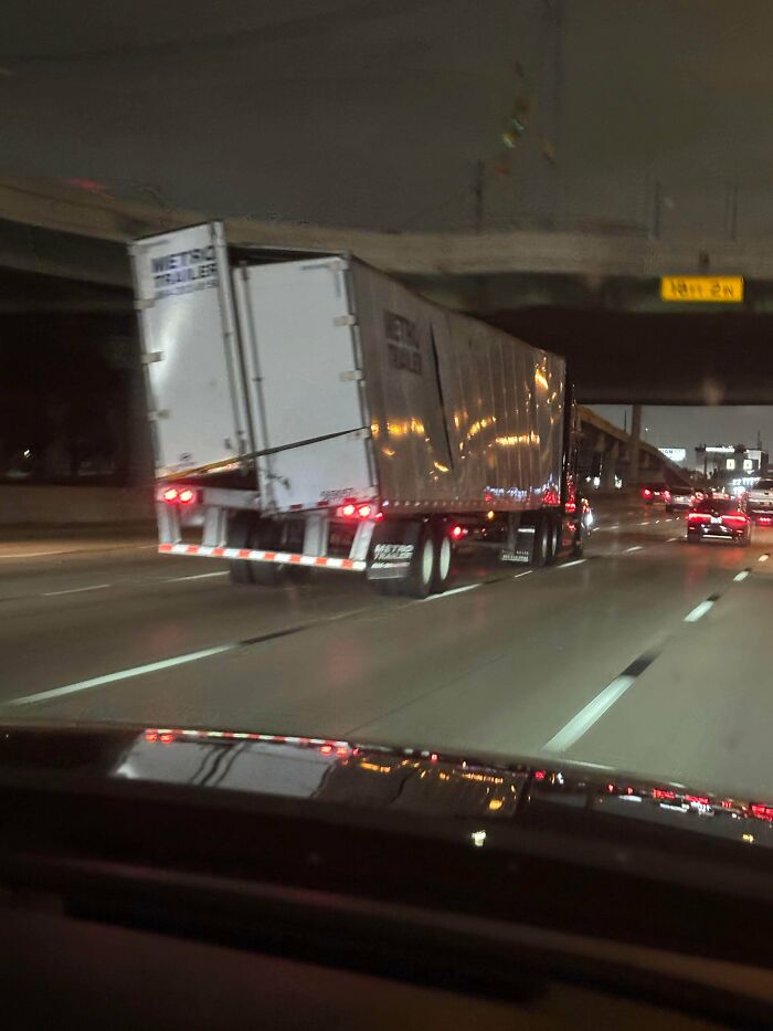A semi-truck pulling a severely damaged and bent cargo trailer on the highway at night.