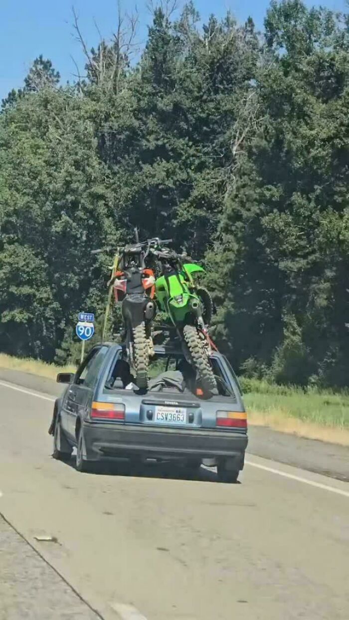 Two dirt bikes stuffed into the hatchback of a small car driving on highway, towing gone wrong example