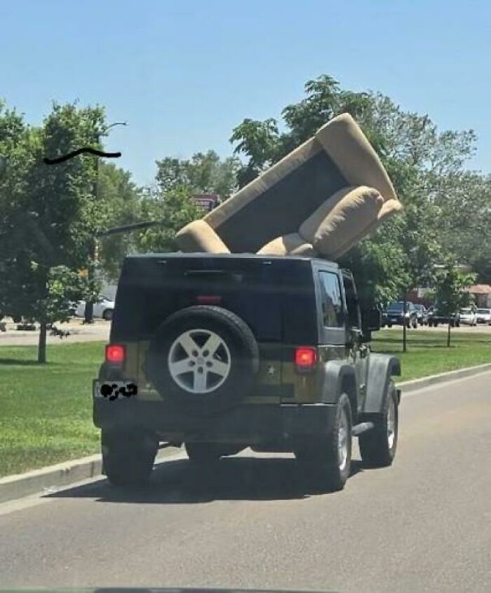 Jeep driving down a street with a large sofa dangerously balanced on its roof, a funny towing gone wrong moment.