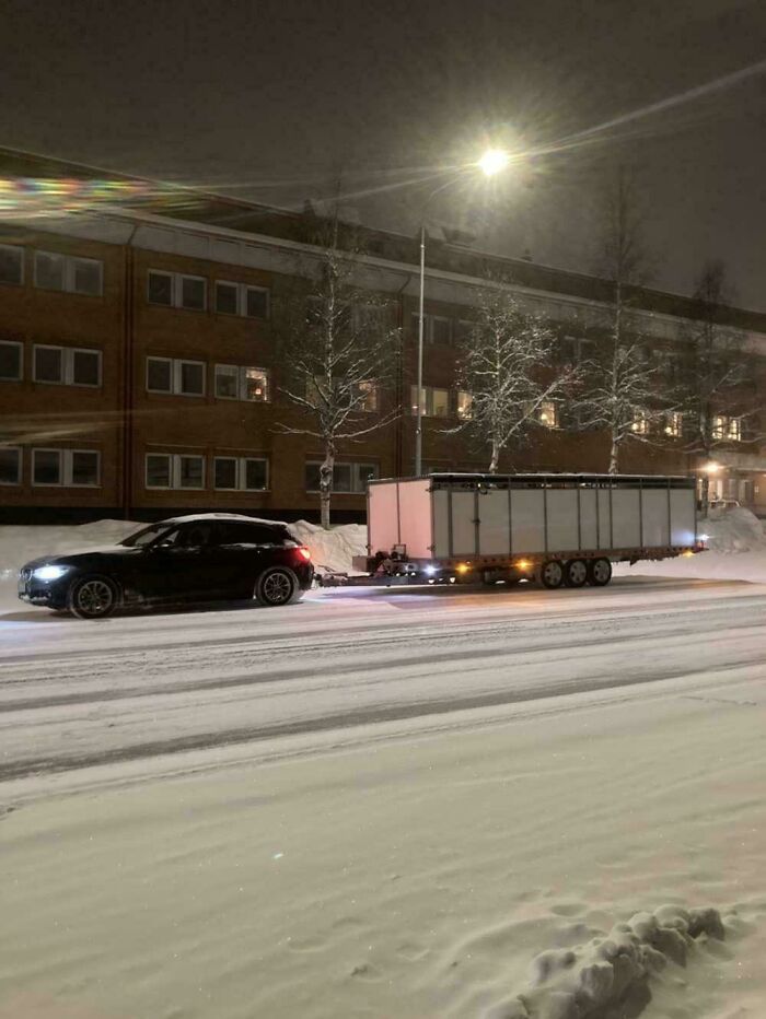 A small hatchback car pulling a massive cargo trailer on a snowy road, demonstrating towing gone wrong.