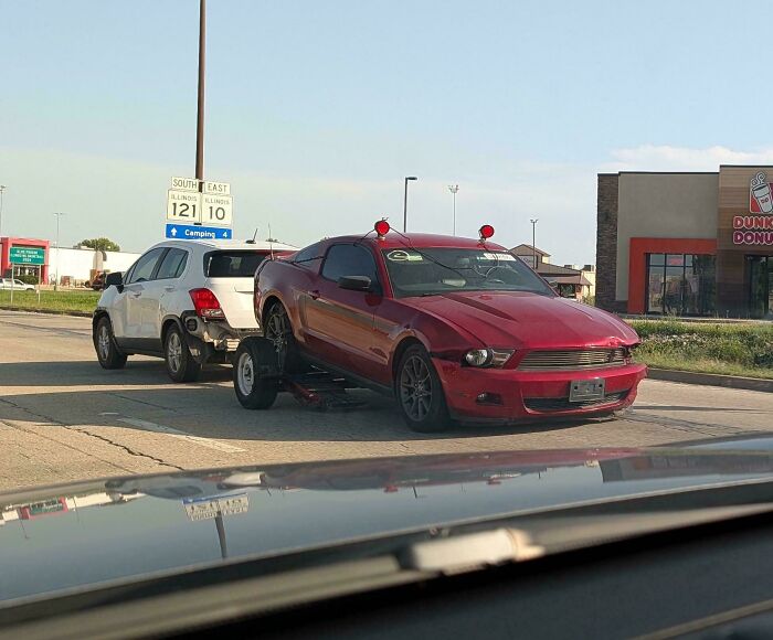 A small white crossover towing a red sports car on a tow dolly in a risky towing setup.