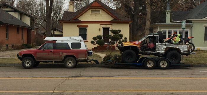 Red SUV towing a trailer carrying a broken truck on residential street, showing towing gone wrong.