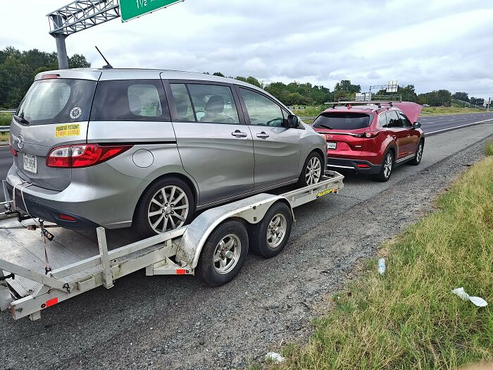 Silver minivan on a trailer pulled by a red car on the side of a highway, showing towing gone wrong.