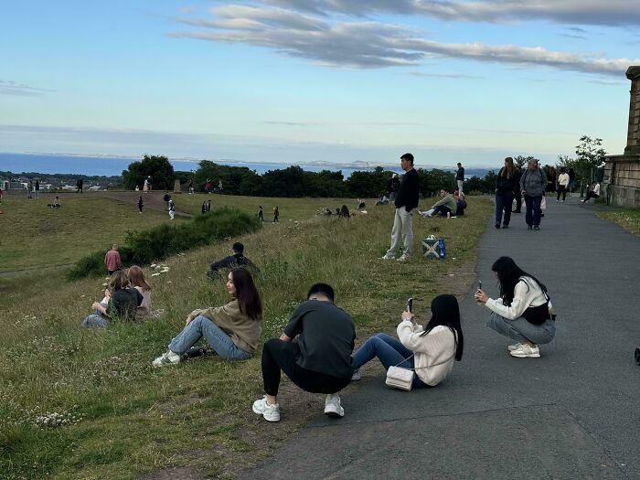 Group of influencers in public spaces taking photos with smartphones on a grassy hill overlooking the sea.