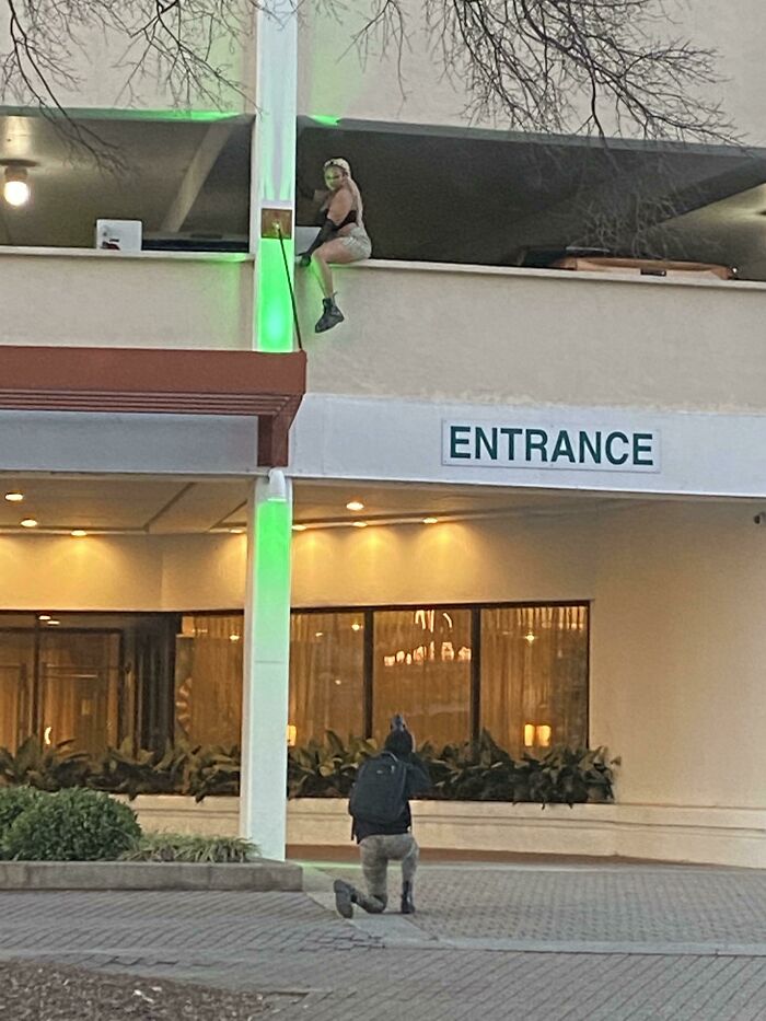 A photographer kneels on a public sidewalk taking pictures of an influencer sitting dangerously on a parking ledge.